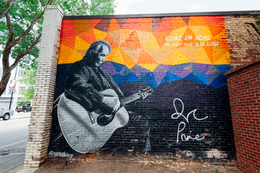 Mural of musician John Prine playing a guitar with the lyrics "Come on home you don't have to be alone"