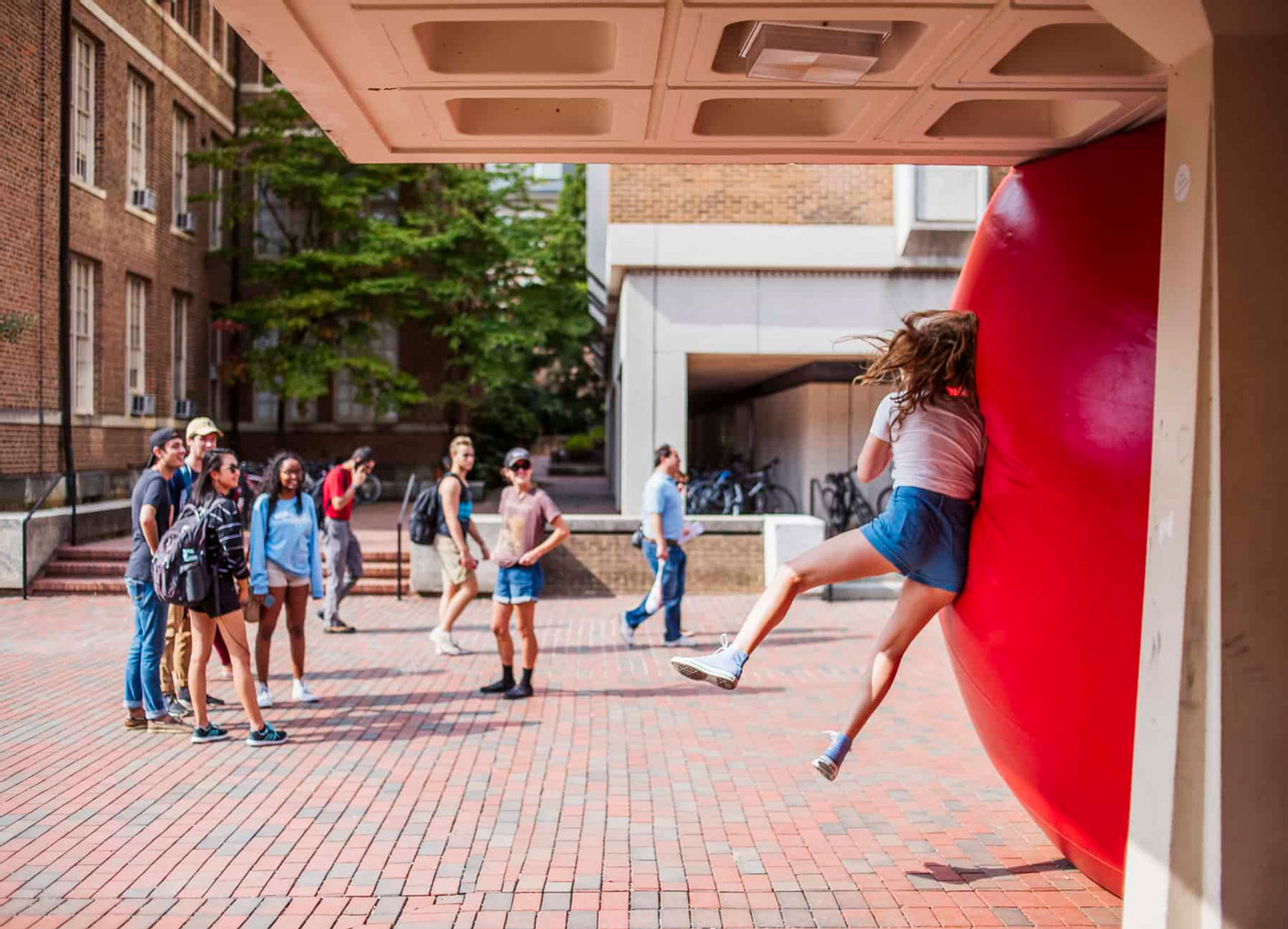 image of large red ball and a girl bouncing against it with a group watching, standing on a brick sidewalk