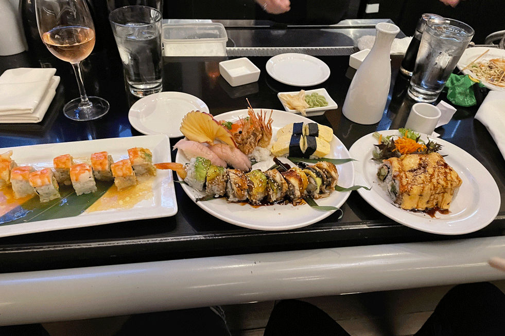 Three plates of sushi sit on a bar. One plate on the left is a roll with shrimp on top, the middle plate has a roll, four pieces of nigiri, the right plate has a roll.